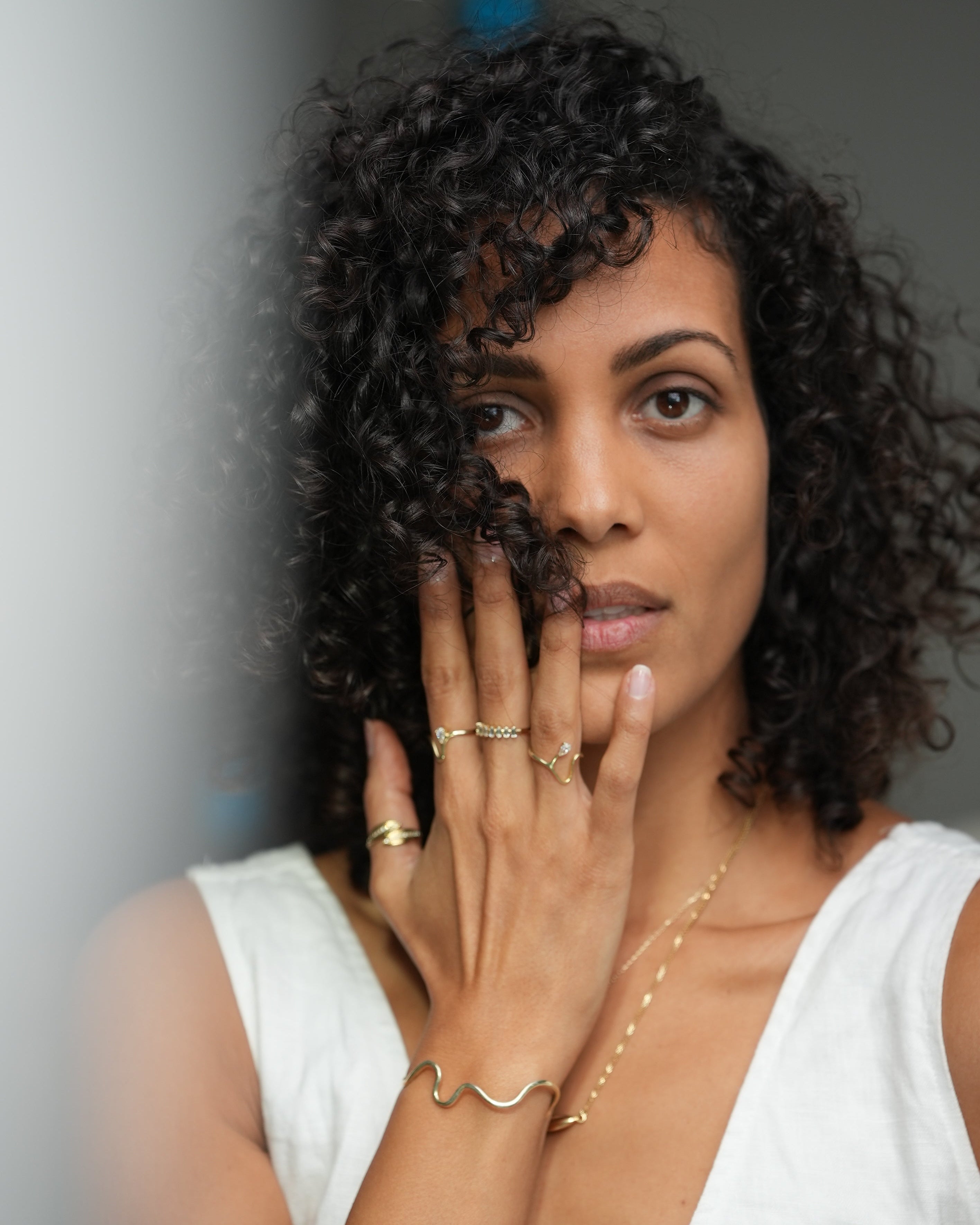Woman with curly hair covering her face with one hand, wearing a white top and gold jewelry.