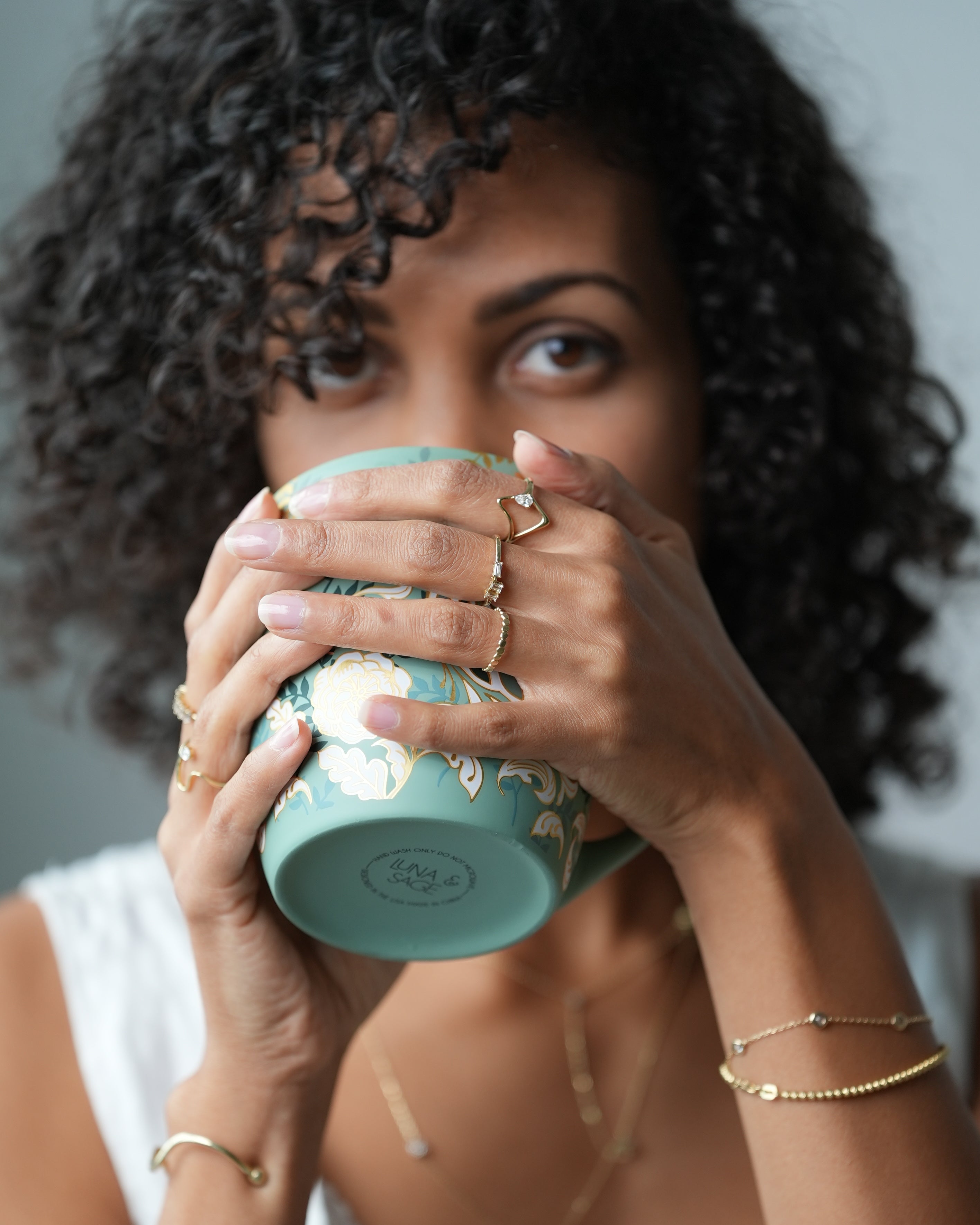 Woman holding a teal mug with a white interior, wearing gold bracelets and rings.