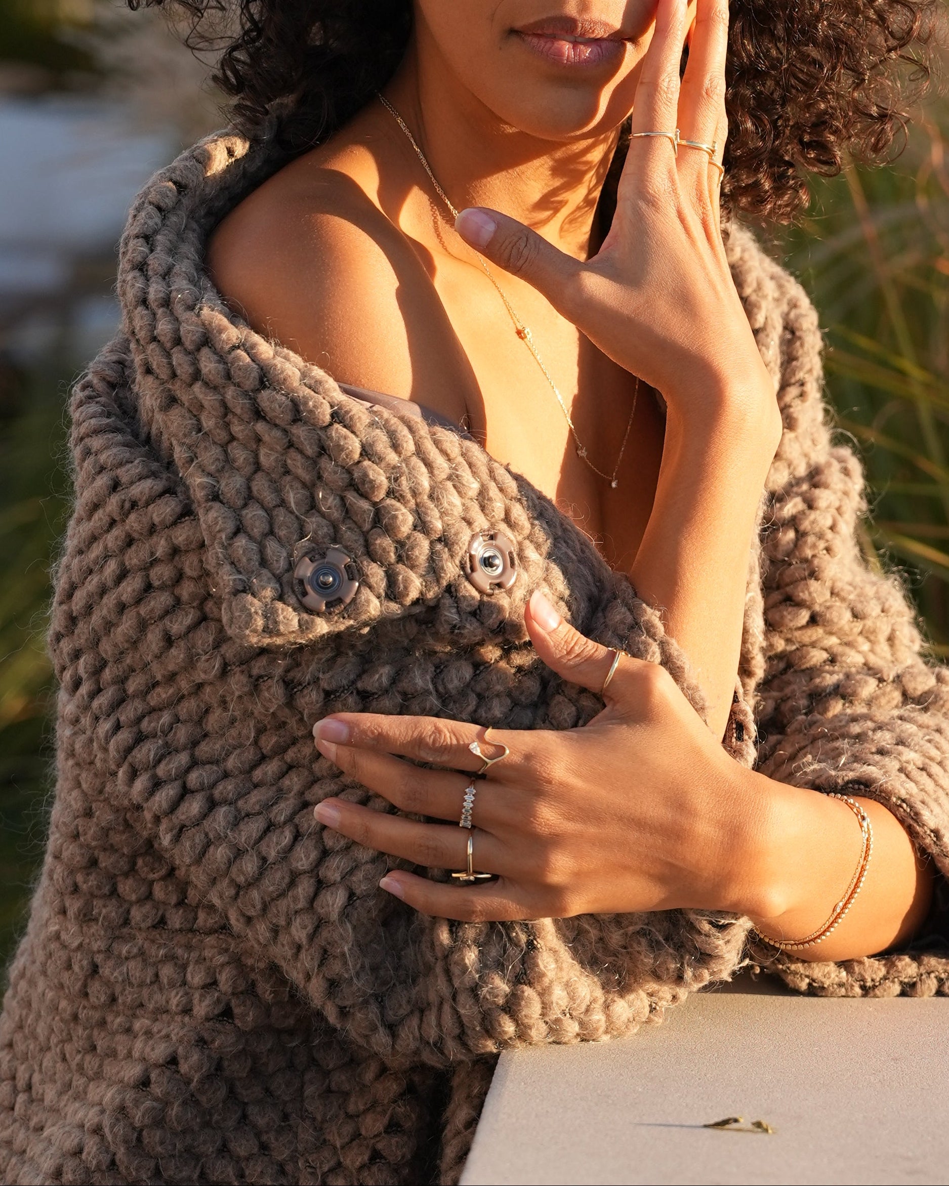Woman wearing a textured brown sweater in an outdoor setting with plants and light background